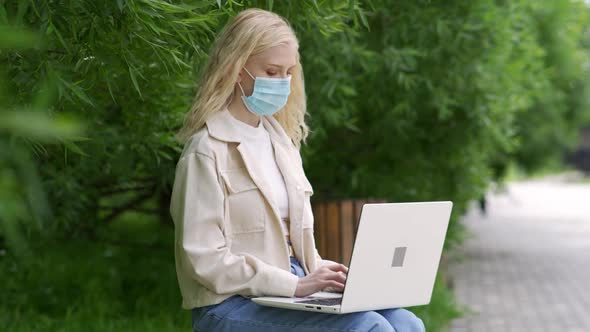 Girl in Antiviral Mask Sits on a Plaid in the Park and Works at a Laptop