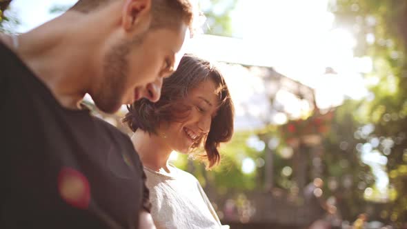 Young Beautiful Couple Smiling Speaking Looking at Tablet Sitting in City Park alt