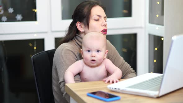 Young Woman with Toddler in Arms Typing on Laptop Remote Work with a Baby at Home