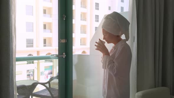 Young Woman Standing at the Window on Sunrise with a Cup of Hot Drink Coffee