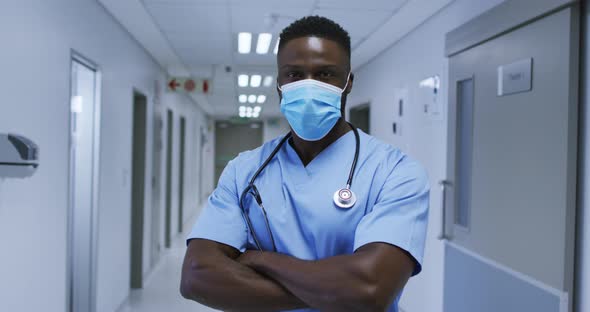 Portrait of african american male doctor wearing face mask and scrubs standing in hospital corridor alt