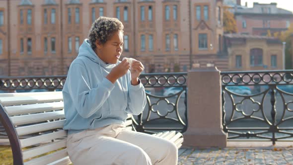 Young Ill African American Coughing and Sneezing in Napkin Outdoor Sitting on Bench. alt
