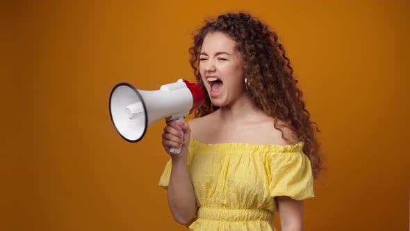 Curlyhaired Young Woman Shouting Into Loudspeaker Against Yellow Background