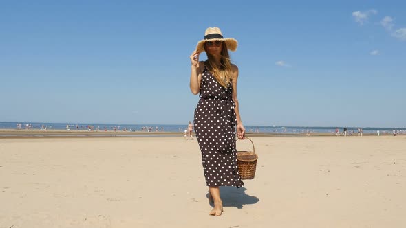 Fashionable Girl Is Walking On The Beach. Young Woman Walks On The Beach With Picnic Basket. 