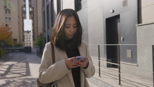 Asian Young Adult Woman Browsing on Her Smartphone While Walking in the City in Autumn alt