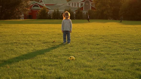 Little Girl Runs with Football Ball and Scores Goal on Green Lawn in Summer Park at Sunset