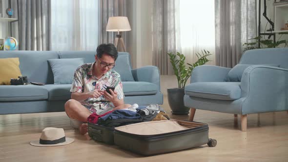 Asian Young Man Using Smart Phone While Packing Clothes In Suitcase At Home, Preparing For Vacation alt
