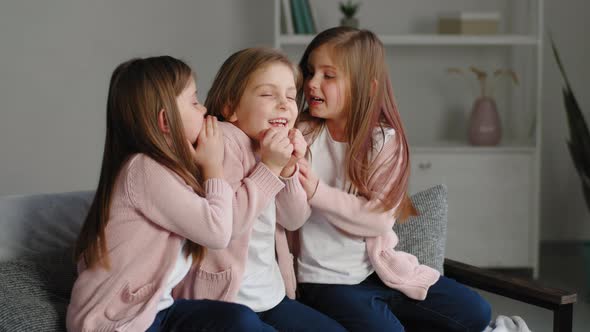 Three Little Girls Sisters in Same Clothes Sitting on Couch at Home Telling Secrets to Each Other alt