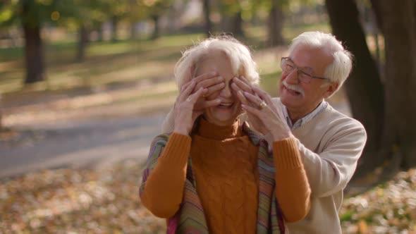 Handsome senior couple embracing in autumn park and man covering eyes of his wife as a surprise