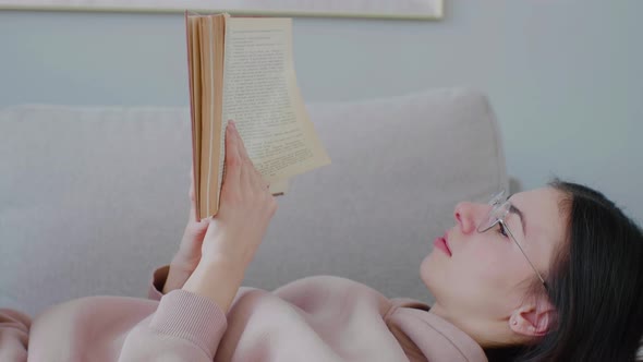 Close up portrait young caucasian woman with glasses reading book lying on sofa.