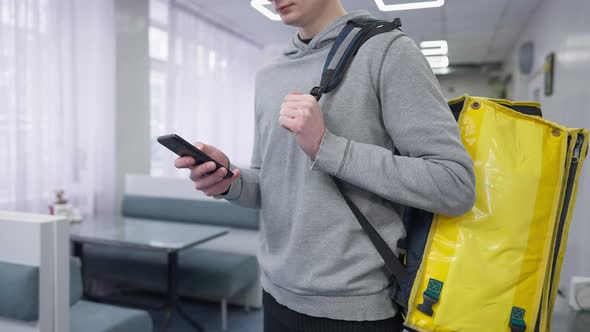 Unrecognizable Male Courier Checking Order in Smartphone App Standing in Cafe with Yellow Thermal alt