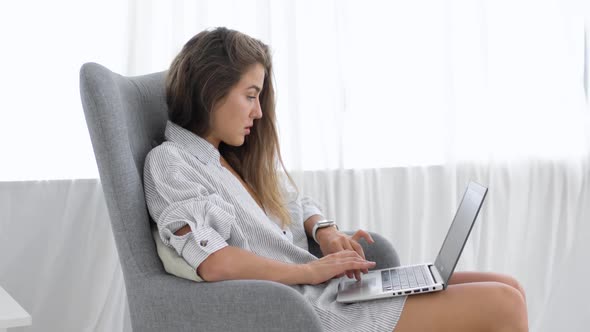 Woman Freelancer Is Working on Laptop Sitting in Armchair at Home, Side View
