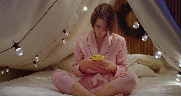 Young Woman Sitting Crosslegged with Smartphone Indoors on Cozy Bed