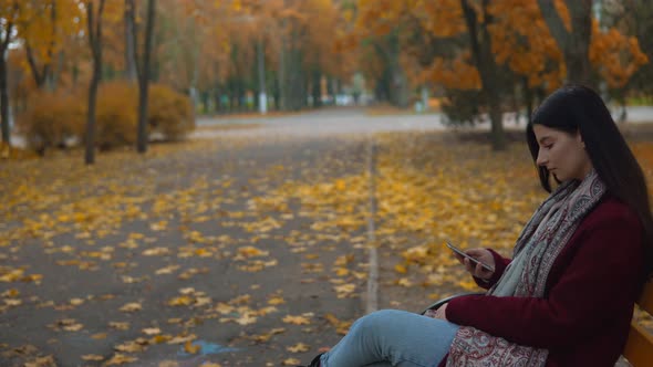 Attractive Woman Walking in Autumn Park Sitting on Bench Using Smartphone