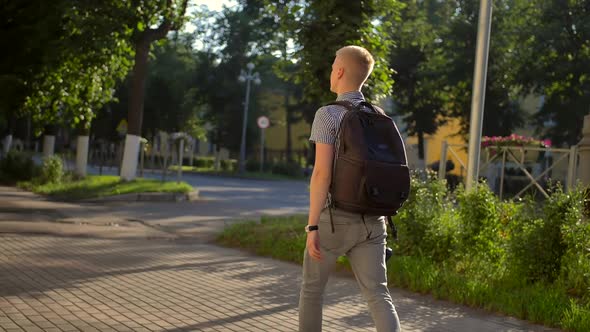 Back-side View of Student Photographer with a Black Backpack Is Walking Along a Town Street alt
