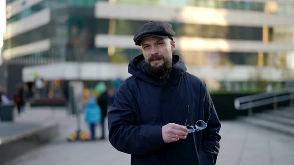 a Mustachioed and Bearded Man in a Cap Takes Off Sunglasses and Walks Against the Background of a alt