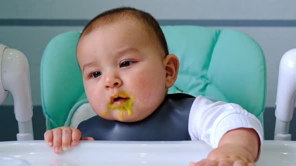 A dirty, joyful baby at the table on a feeding chair got his mouth dirty in vegetable puree and knoc alt