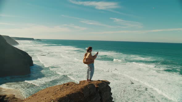 Woman Stand on Edge of Ocean Cliff with Phone alt