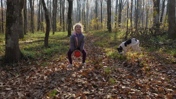 A blond child playing with a ball and a dog in an autumn forest alt