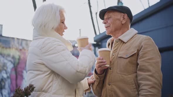 An Elderly Couple Drinks From Coffee Cups on a Sunny Street Talking and Smiling