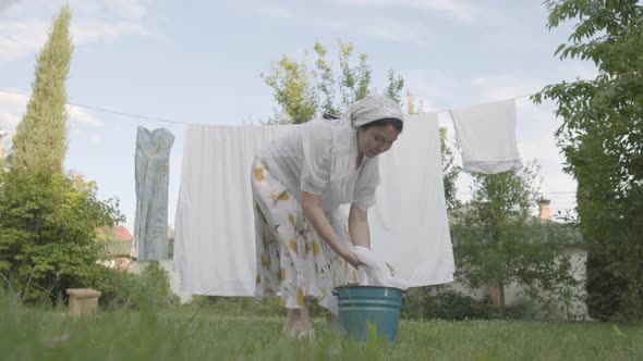 Attractive Senior Woman with a White Shawl on Her Head Hanging Bed Linen on the Rope in the Garden alt