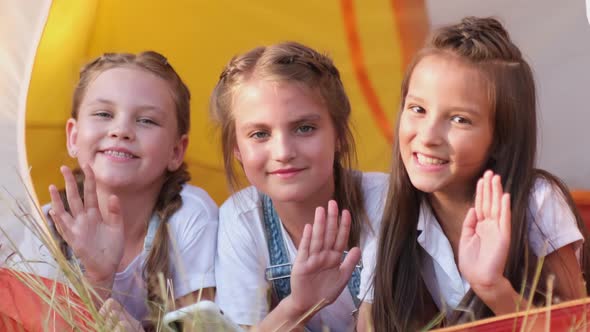 Three Little Girls are Sitting Near a Tent in the Woods They are Waving Their Hands at the Camera alt