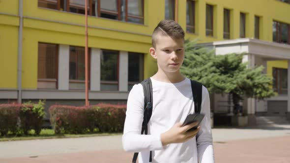 A Caucasian Teenage Boy Takes Selfies with a Smartphone  a School in the Background