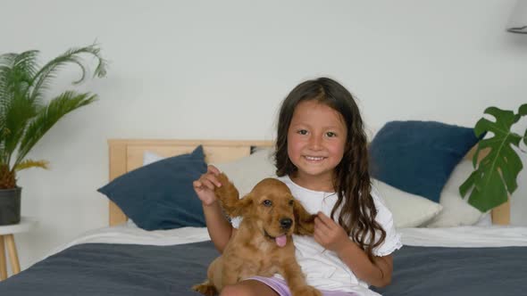 Little cute hispanic girl playing on bed and having fun with cocker spaniel puppy.