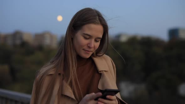 Woman Is Chatting in the Smartphone Against the Background of the Evening City
