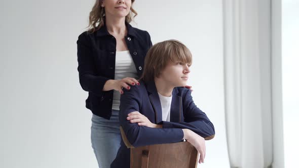 A Mother with Son in Blue Clothes Poses on a Chair By Window for a Photo Shoot alt