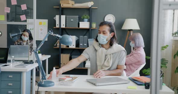 Young Man Wearing Mask Coming to Office Greeting Colleagues Spraying Hands with Sanitizer alt