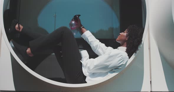 Young black woman is sitting on window and spraying herself with water to cool down in a summer day alt