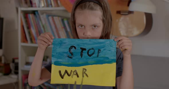 Child shows a poster Stop war inscription on Ukranian flag paint in his home. alt
