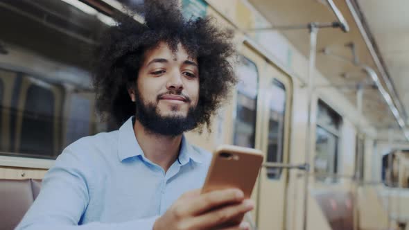 Stylish Expressive Curly Dark-Skinned Man Sitting in Subway Wagon with Smartphone. Riding at Public alt