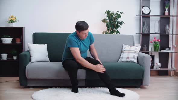 A Caucasian Man with a Painful Knee Injury Is Sitting on the Couch and Will Rupture His Knee a