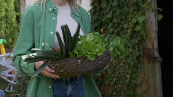 Stylish woman in green shirt with basket full of vegetables stands on ivy background alt