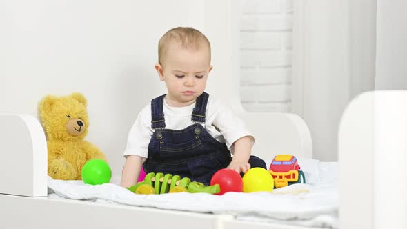 Baby Boy in Denim Overalls Sitting on the Bed Plays Toys and Throws Plastic Colored Balls alt
