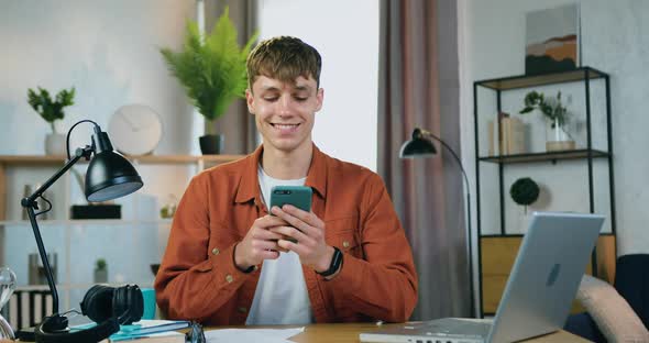 Young Guy Sitting at the Table at Home and Uses His Mobile to write Message to His Mates alt