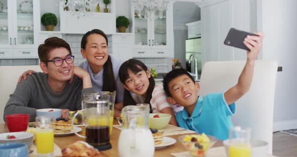 Happy asian parents in kitchen having breakfast with son and daughter, son taking group selfie alt