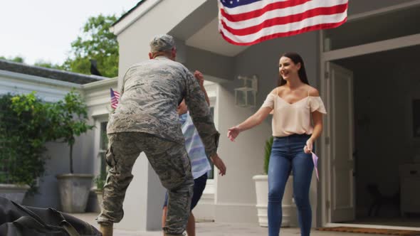 Happy caucasian male soldier lifting son and greeting wife with flag hanging outside their house alt