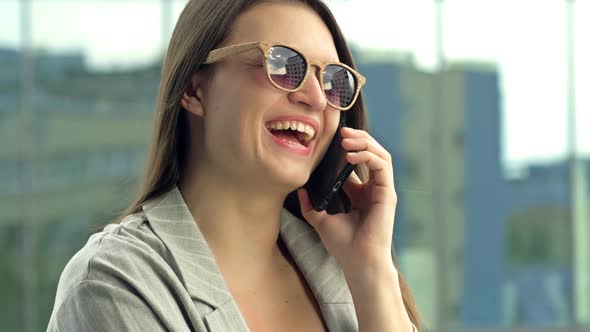 Cheerful Young Woman in Sunglasses Talking on the Phone While Standing on a City Street alt