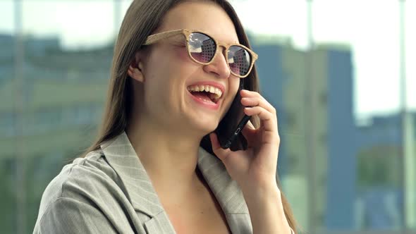 Cheerful Young Woman in Sunglasses Talking on the Phone While Standing on a City Street