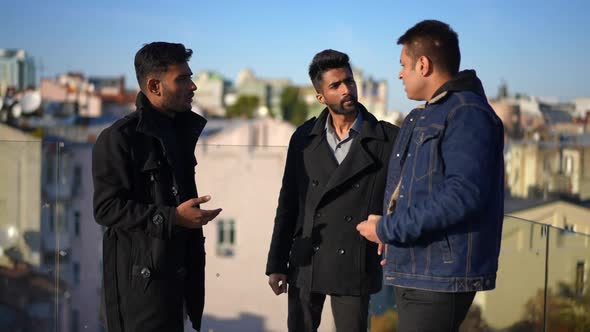 Three Positive Male Friends Talking Standing at Glass Fence on Rooftop alt
