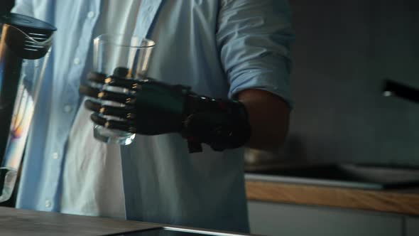 Male person with black artificial hand prothesis pours water into transparent glass alt