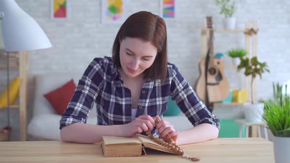 Portrait Young Woman Sitting at a Table with a Rosary in Her Hands and Reading a Book alt