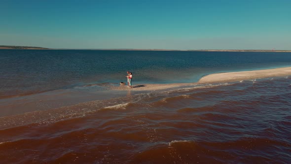 Aerial View From a Drone Flying Around of Happy Couple and Dog Walking By Sand Beach Between Water alt