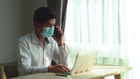 Young man in mask taking a call on his mobile phone checking information received on his laptop  alt