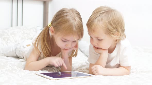Little Caucasian Kids Use Tablet Sitting on Bed alt