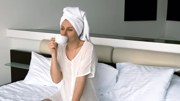 Girl Wearing Bathrobe and Towel Drinks Coffee While Sitting on the Bed Looking at the Camera