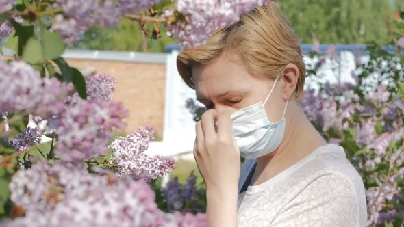 A Young Beautiful Woman in a Medical Mask Sniffs Lilac Flowers